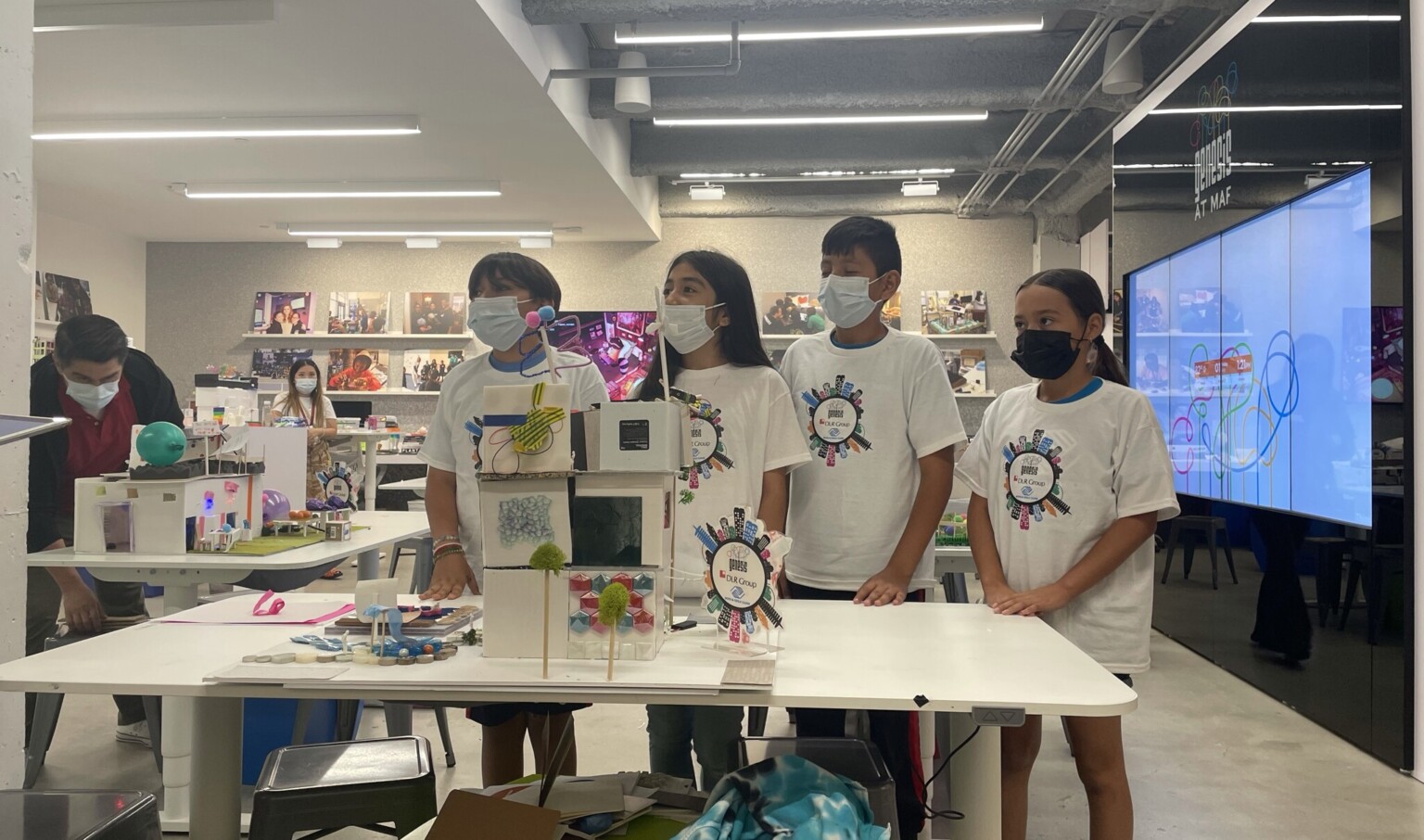 four students in masks standing at a table with an architecture model in front of them, large tv on the far wall four students in masks standing at a table with an architecture model in front of them, large tv on the far wall