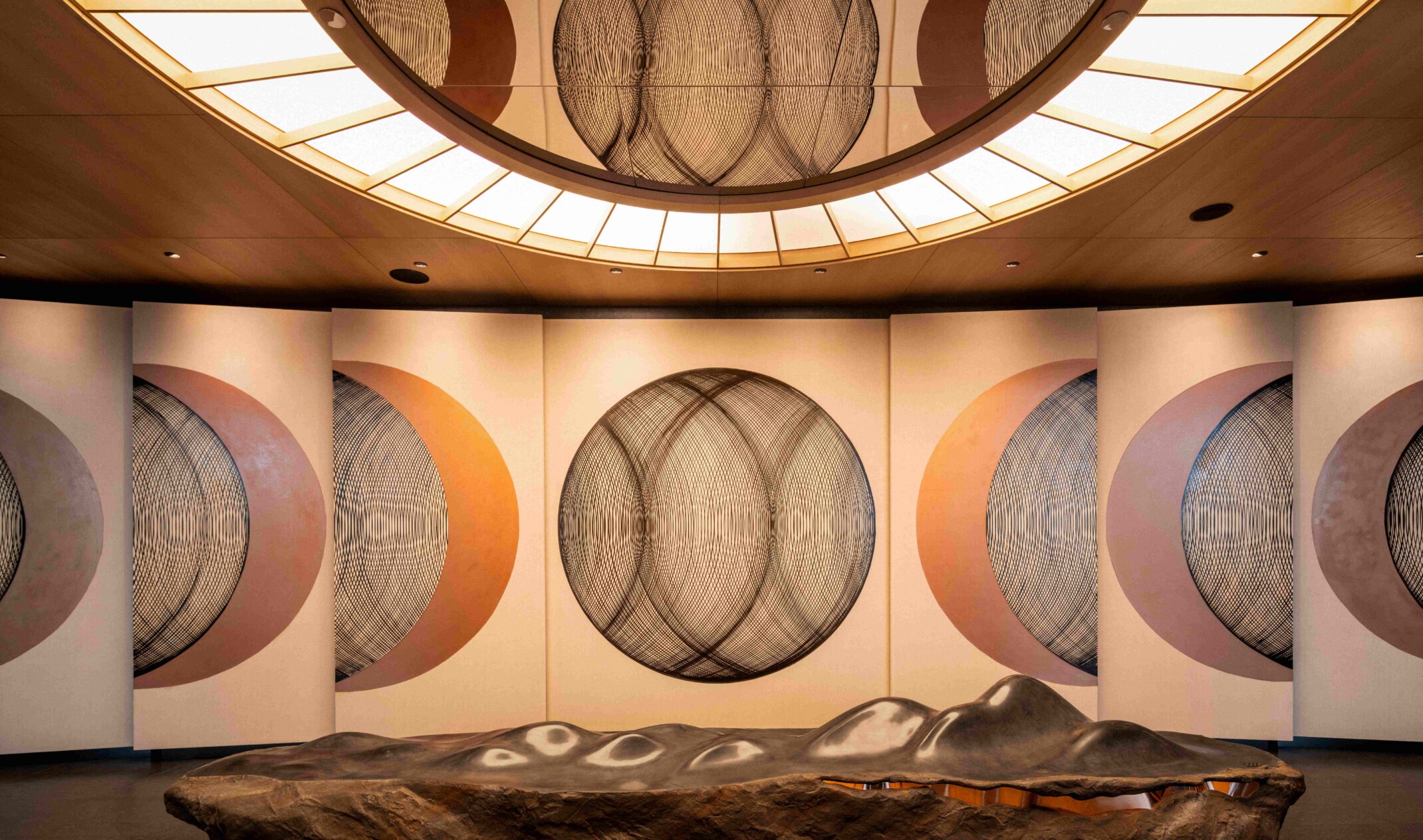 Circular geometric space with ceiling mirrors and lighting highlighting the artwork; rock-like bench in the middle of the room