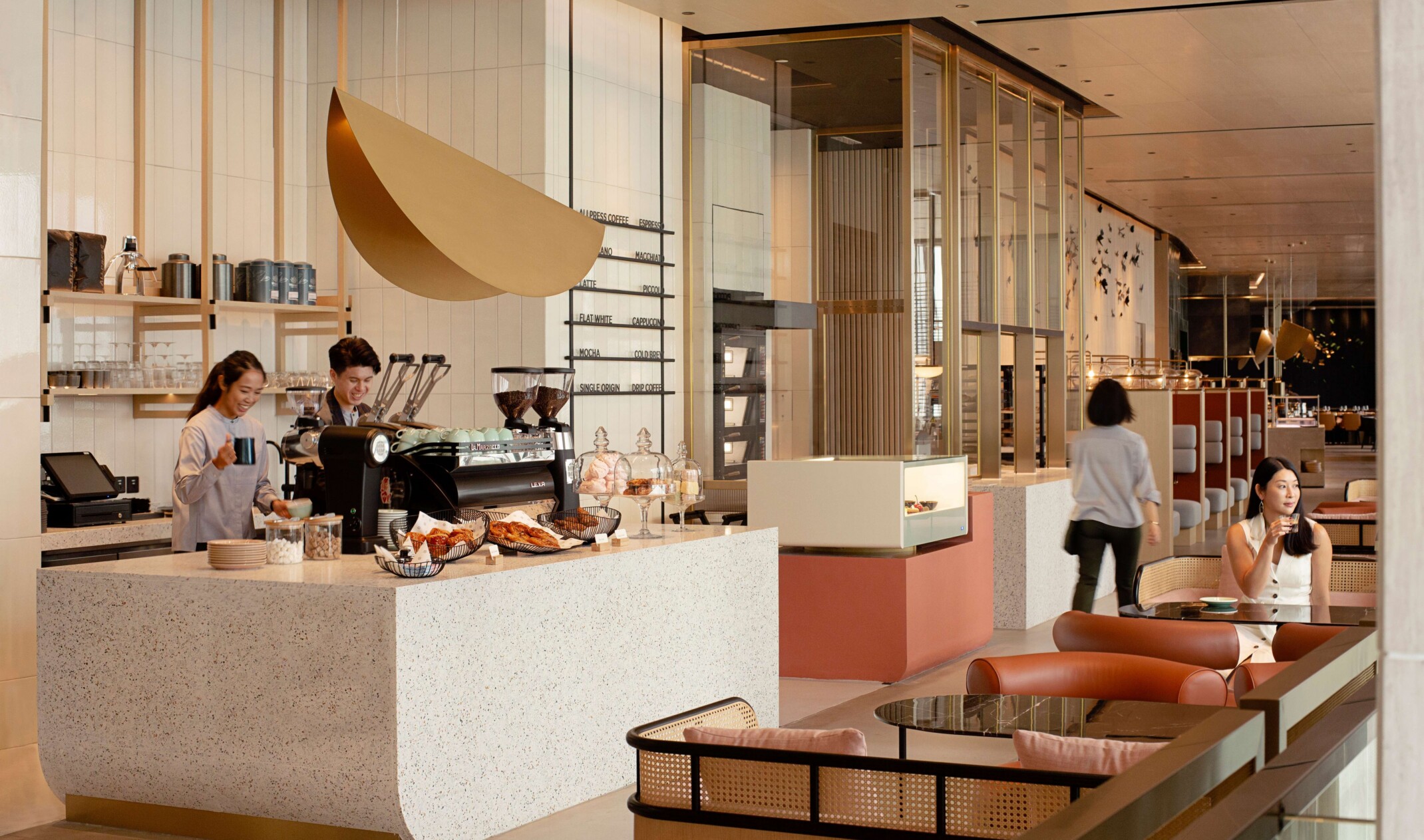 light wood and pastel cafe area; foreground booths with a woman drinking coffee; barista and pastry counter in the background