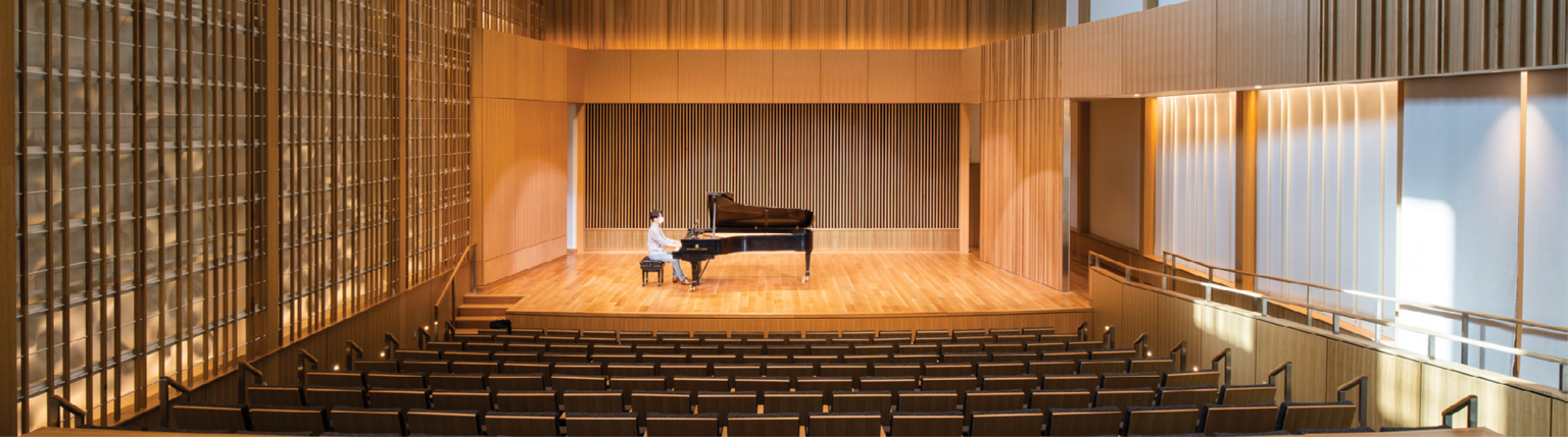 Man sitting at piano in double height recital room auditorium with theater seating. Wood panel designs on walls and wood acoustic panel