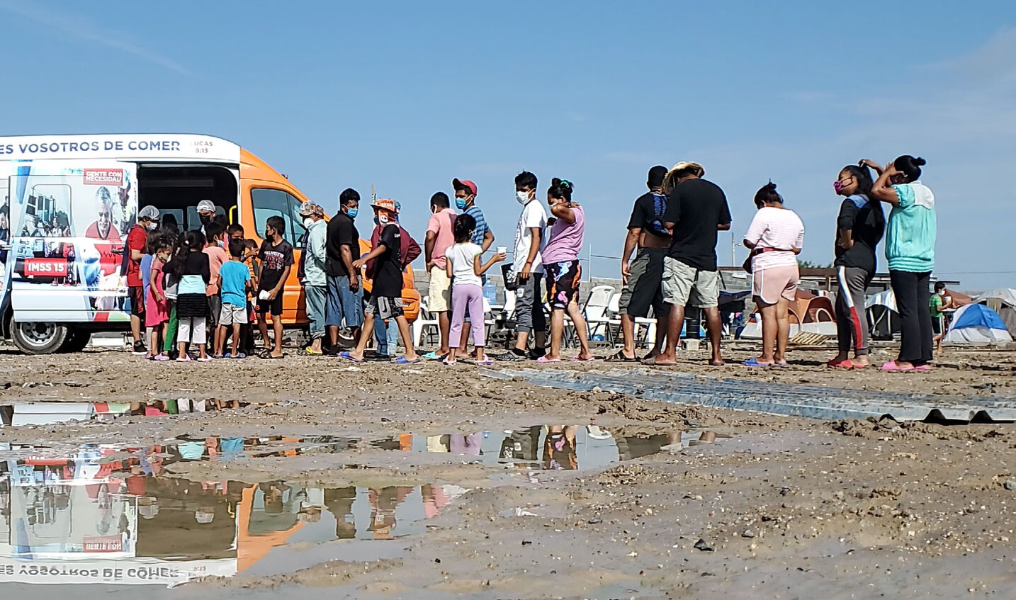 Line of people stand in muddy open space. Line leads to food donation van with side doors opened. Tents visible behind Line of people stand in muddy open space. Line leads to food donation van with side doors opened. Tents visible behind