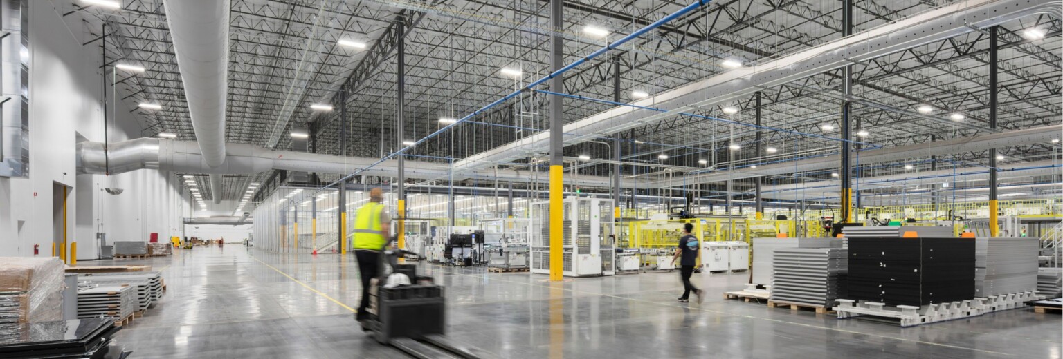 Worker driving a forklift through a warehouse and light manufacturing floor with wide circulation aisles, equipment, and industrial work areas.