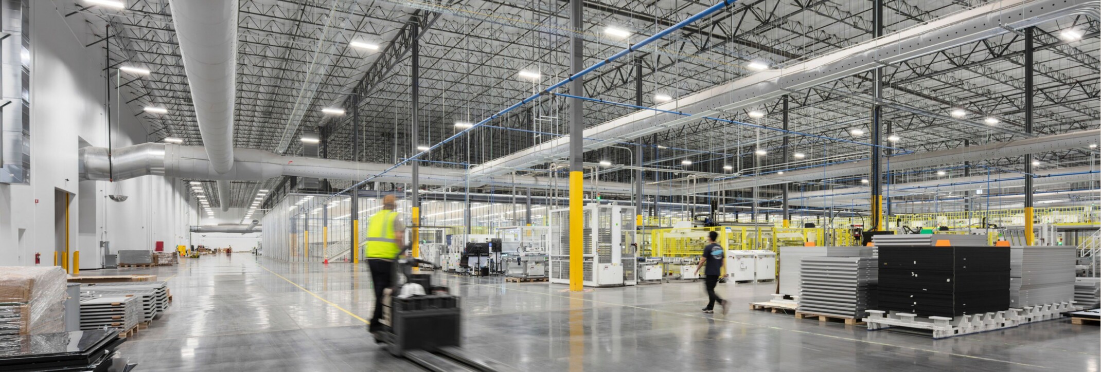 Worker driving a forklift through a warehouse and light manufacturing floor with wide circulation aisles, equipment, and industrial work areas.