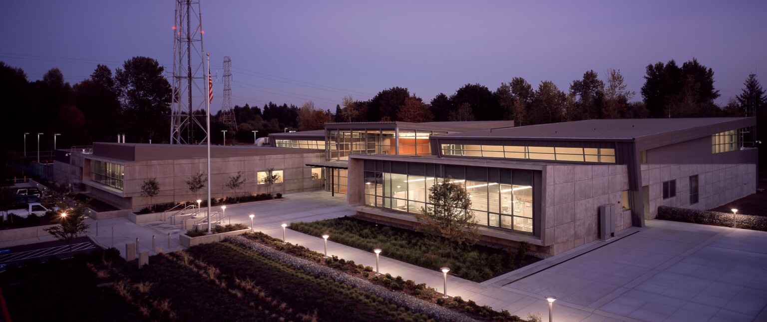 Angular single story two tone grey building with large windows illuminated from within and along pathway at night