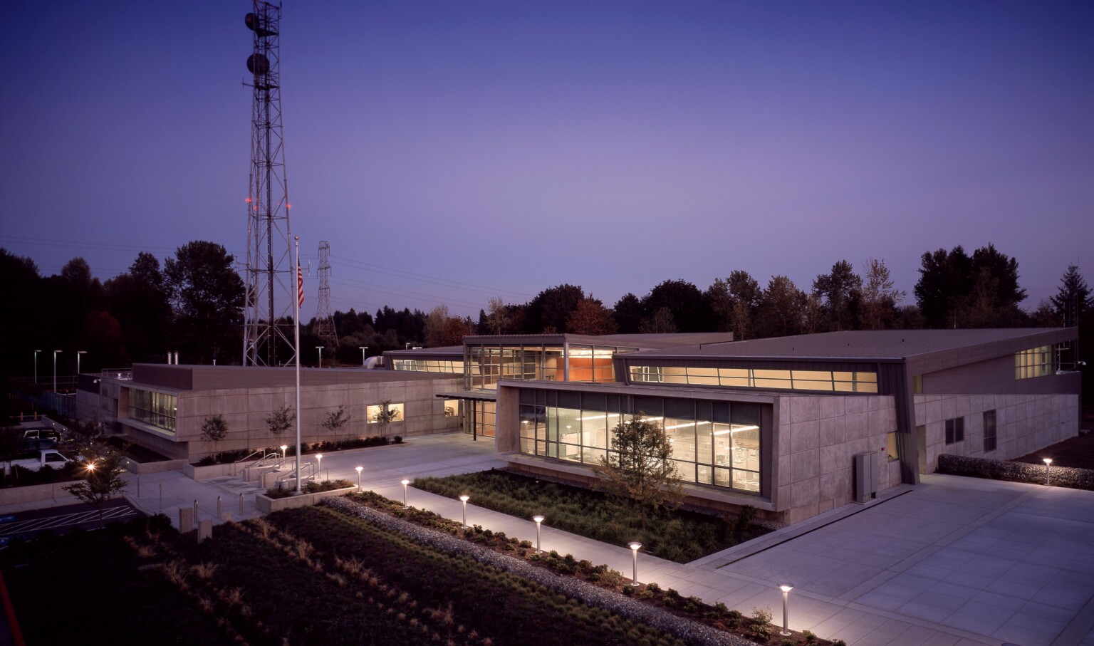 Angular single story two tone grey building with large windows illuminated from within and along pathway at night