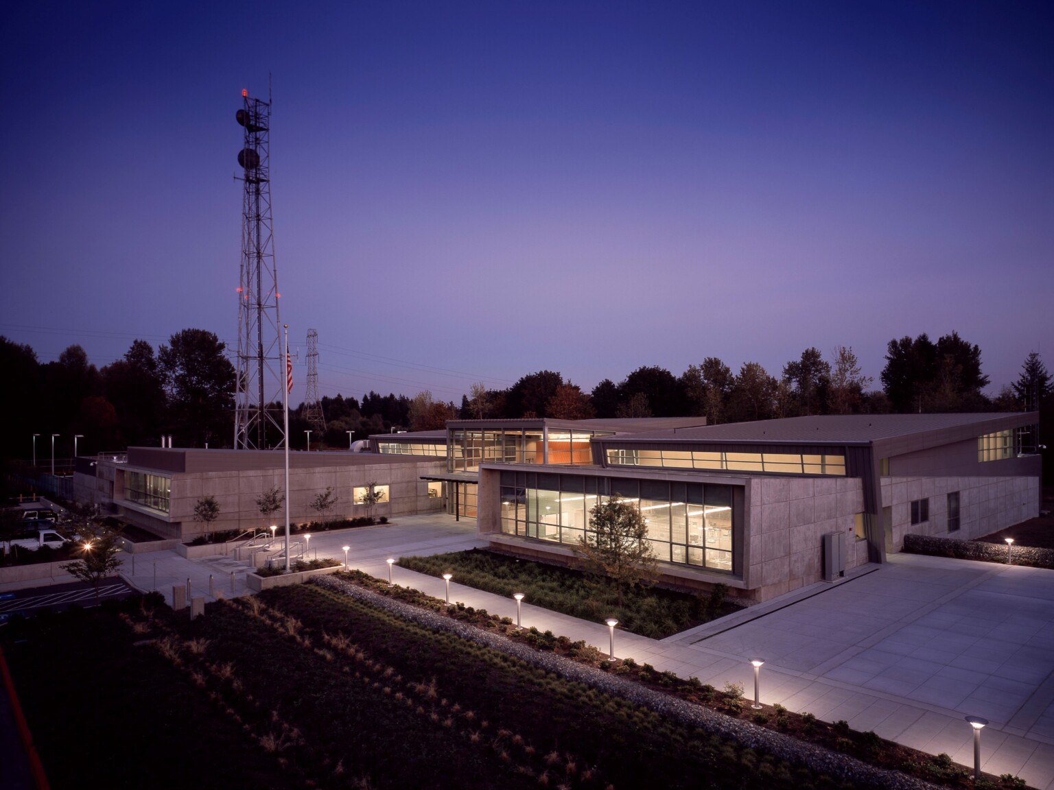 Angular single story two tone grey building with large windows illuminated from within and along pathway at night