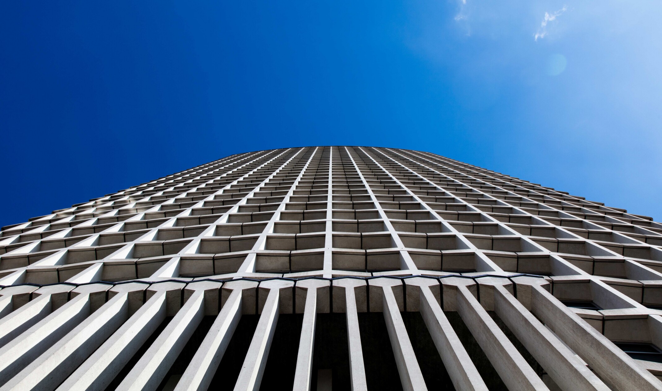 Ground floor directly up view of a primarily cement skyscraper; tall pillars connect to cubic cutouts on the facade