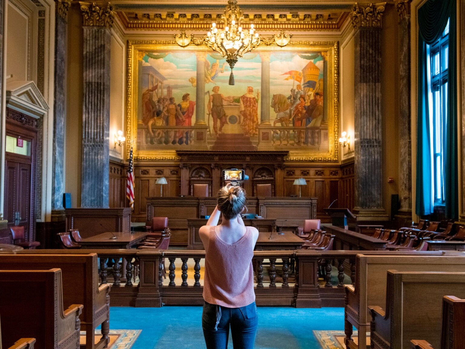 woman taking a video of an historic courtroom, vintage chandelier hanging from ceiling over dark walnut and a large mural painting