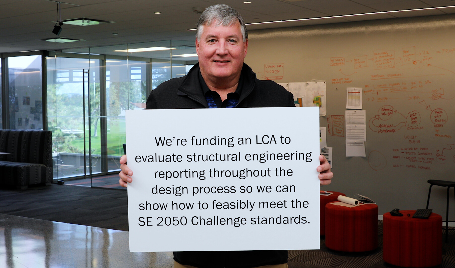 a man in black holding a sign that reads we're funding an LCA to evaluate structural engineering reporting through the design process so we can feasibly meet the SE 2050 Challenge standards.