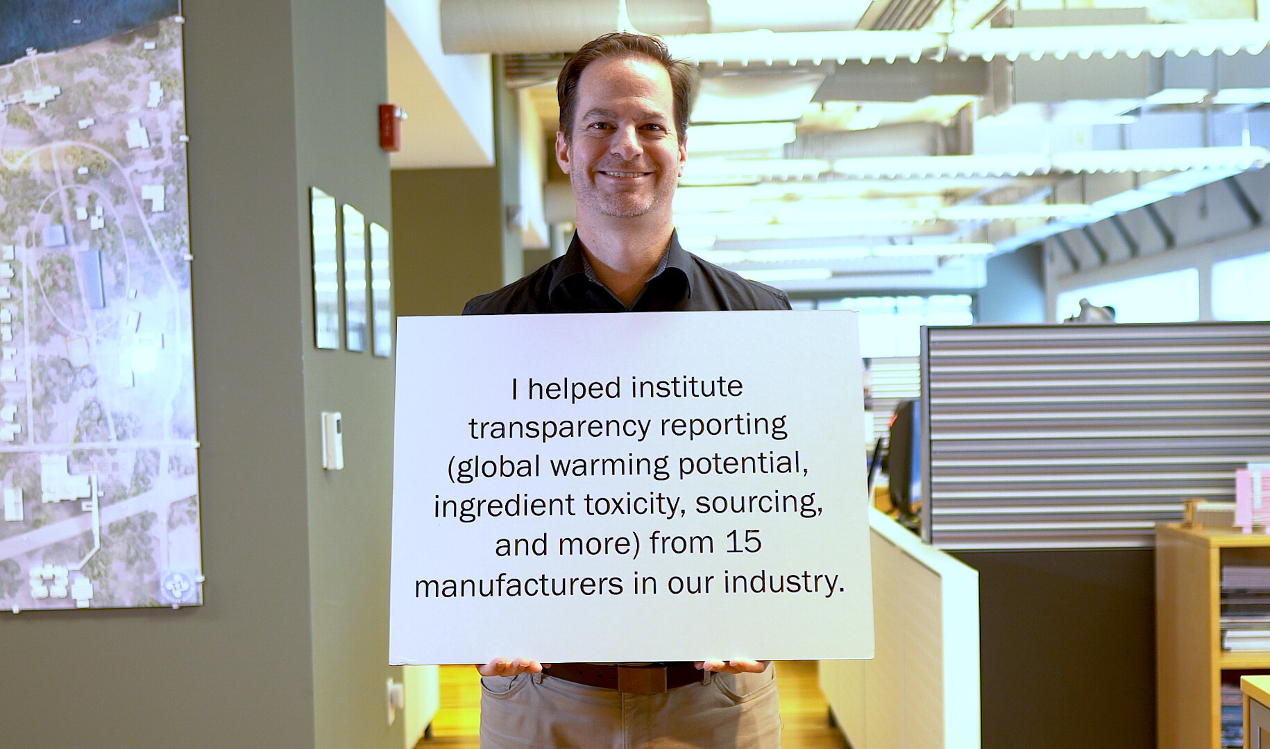 smiling man holds a sign that reads I helped institute transparency reporting global warming potential ingredient toxicity sourcing and more from 15 manufacturers in our industry.