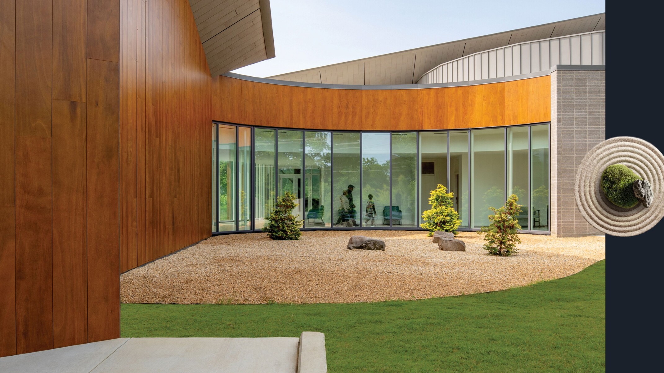 Semicircular exterior courtyard wrapped by the wood and window-clad exterior of the building. The courtyard surface features gravel, small boulders, and low evergreen trees, bounded by lush, short grass; circular sand garden with moss and a rock in the center to the right of the image