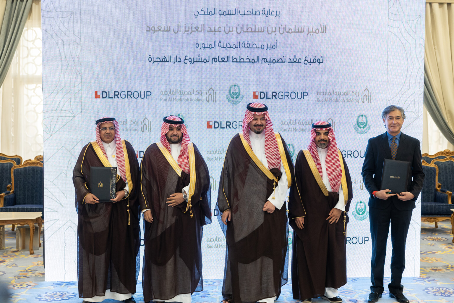 Five men standing in front of a white projection with DLR Group and Rua Al Madinah Holding across the background; two men on the ends holding portfolios with contracts Five men standing in front of a white projection with DLR Group and Rua Al Madinah Holding across the background; two men on the ends holding portfolios with contracts
