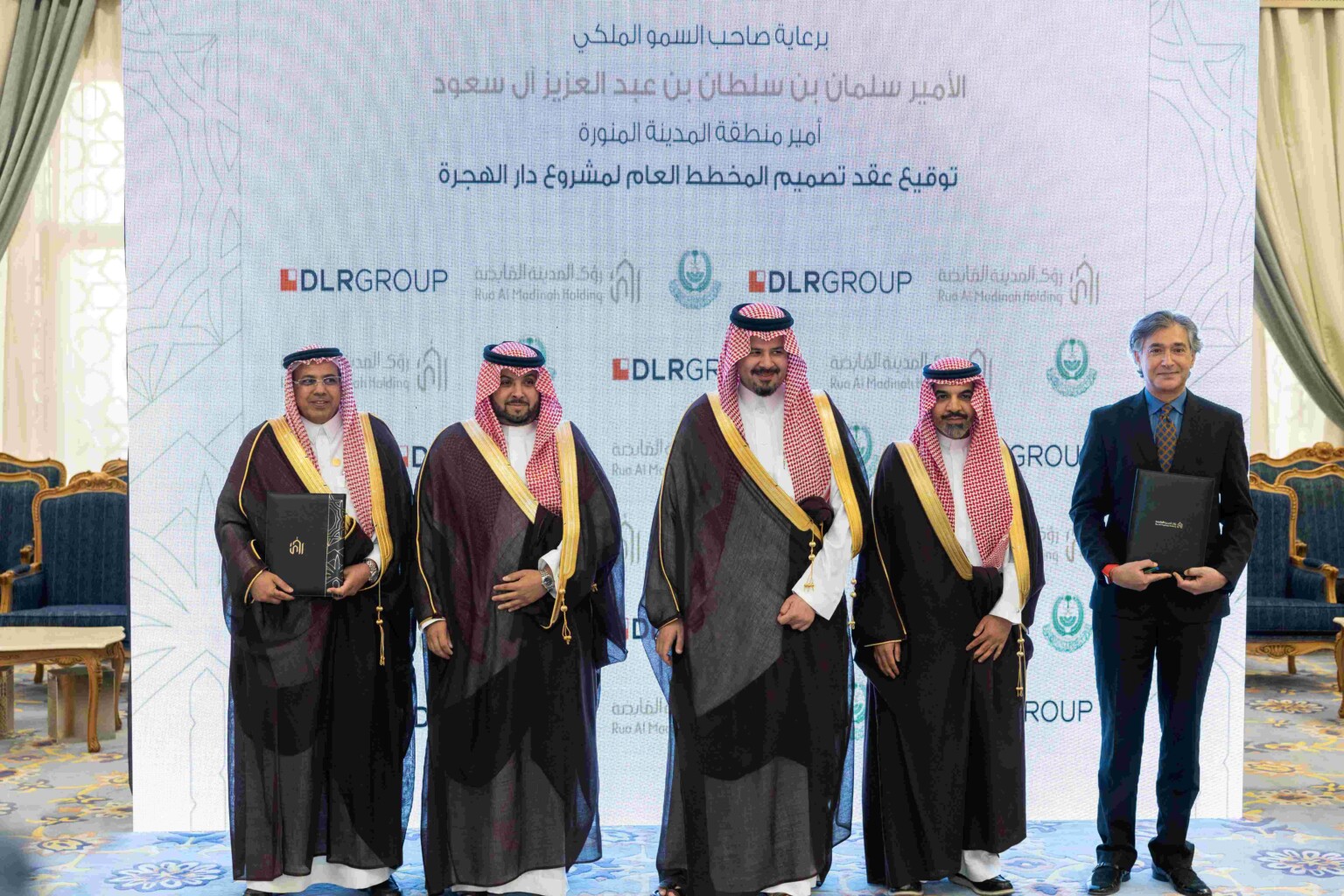 Five men standing in front of a white projection with DLR Group and Rua Al Madinah Holding across the background; two men on the ends holding portfolios with contracts