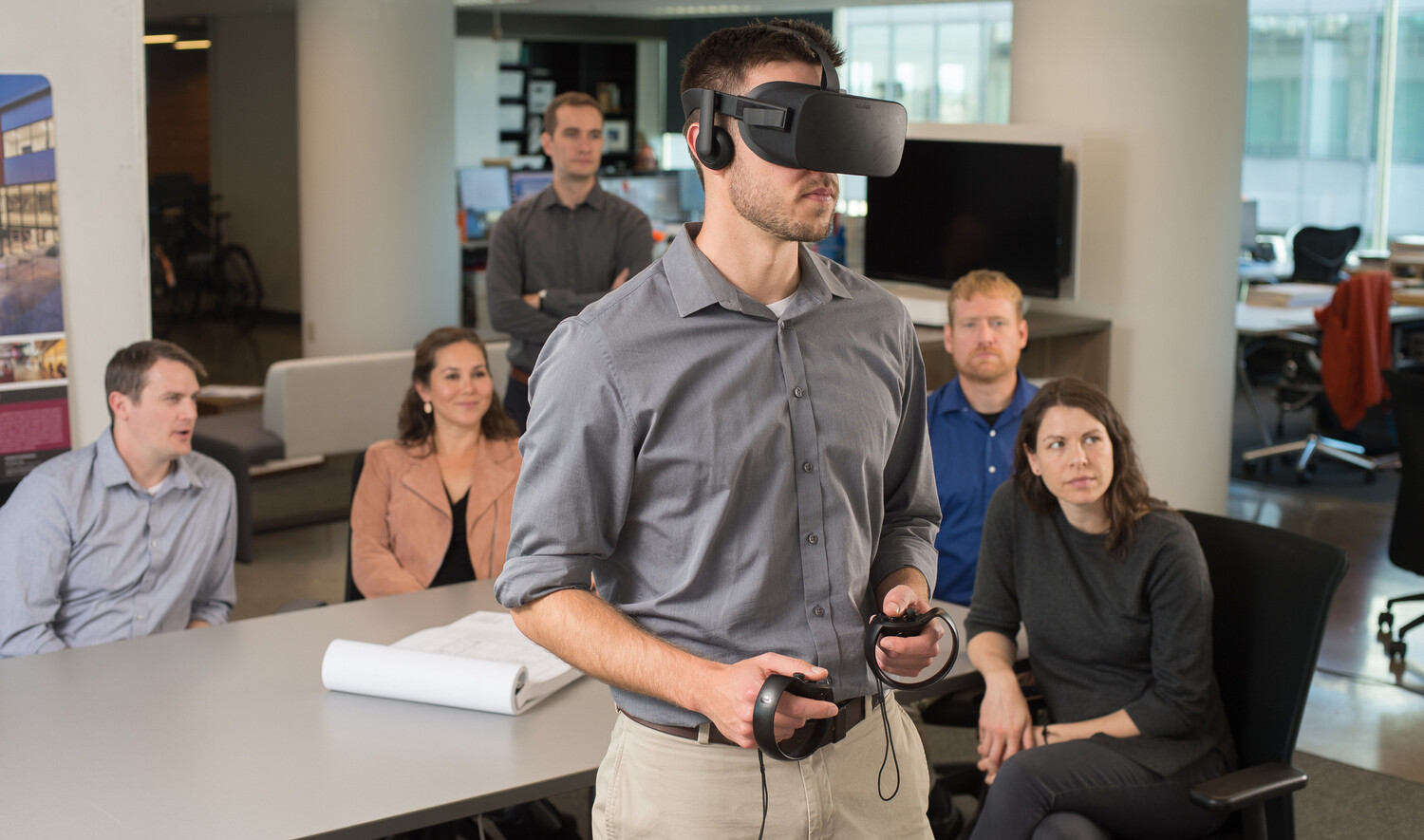 a group of young professionals watches another team member in a virtual reality headset engage in an experience