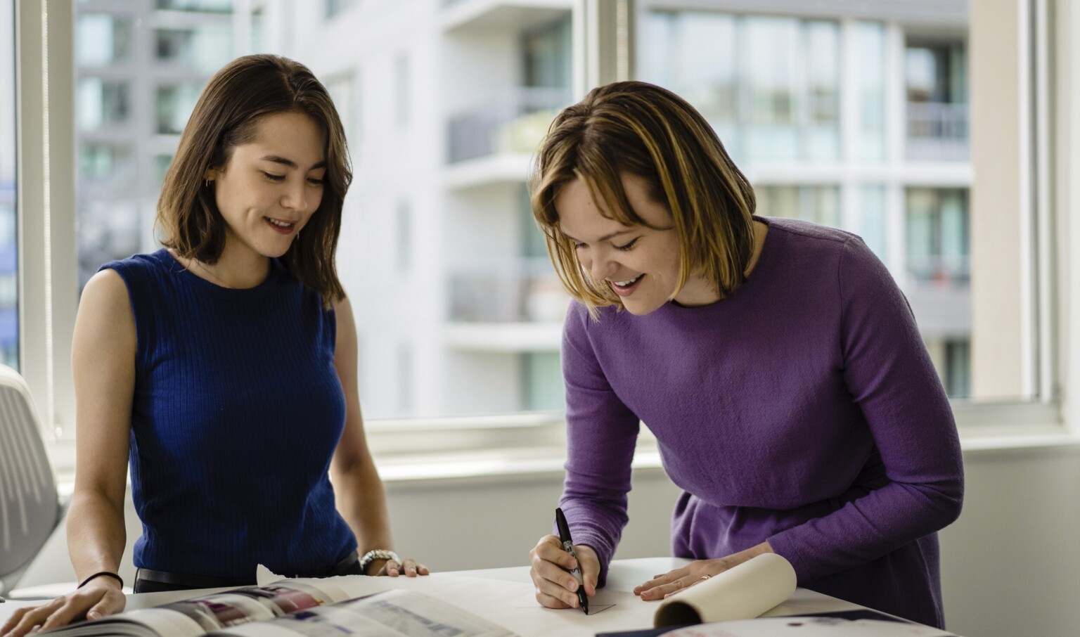 One person wearing blue and one wearing purple reviewing documentation in an office in front of a window One person wearing blue and one wearing purple reviewing documentation in an office in front of a window
