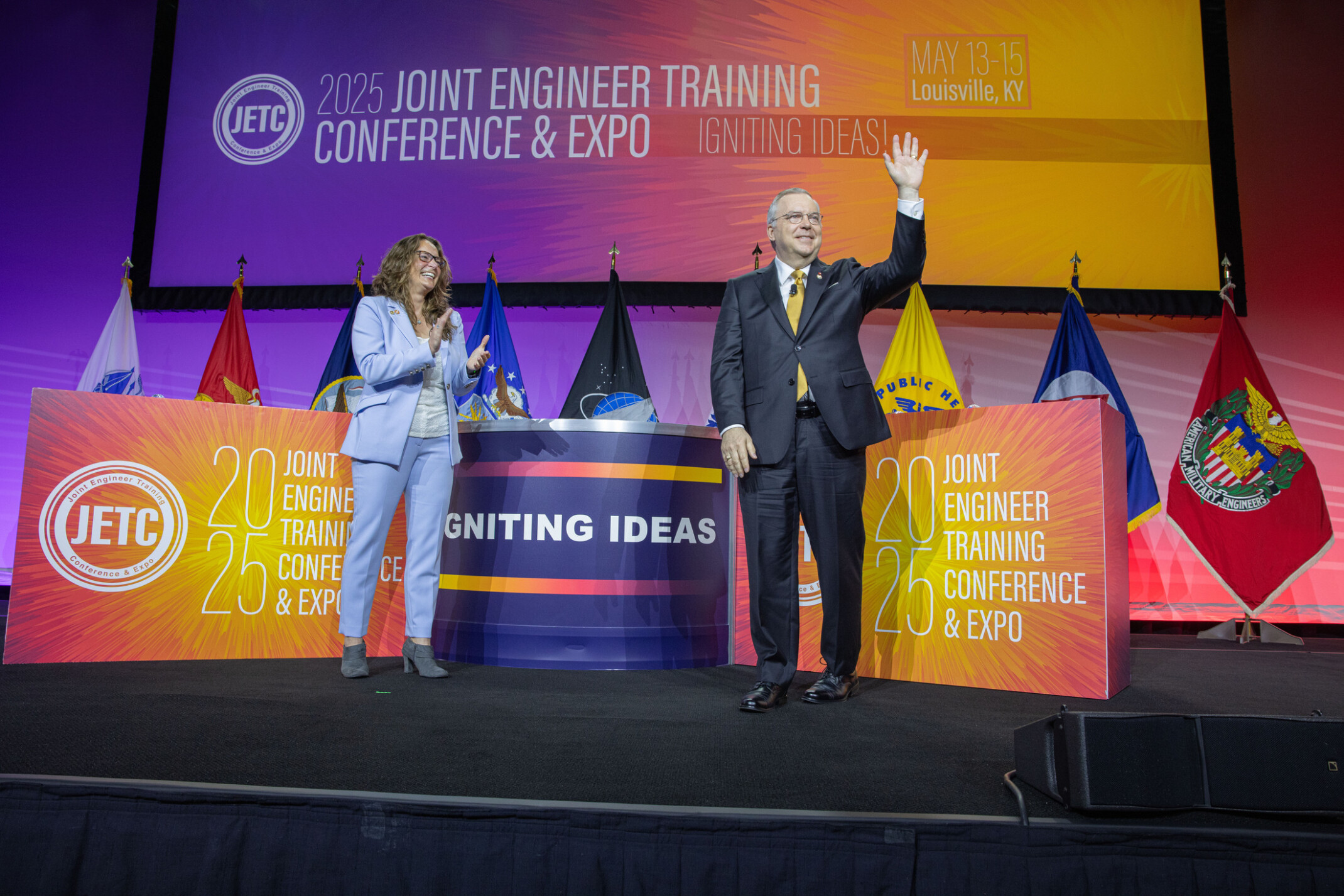 Michael Huffstetler elected to the Society of American Military Engineers; two people on stage celebrating the election with military flags and colorful banners behind them