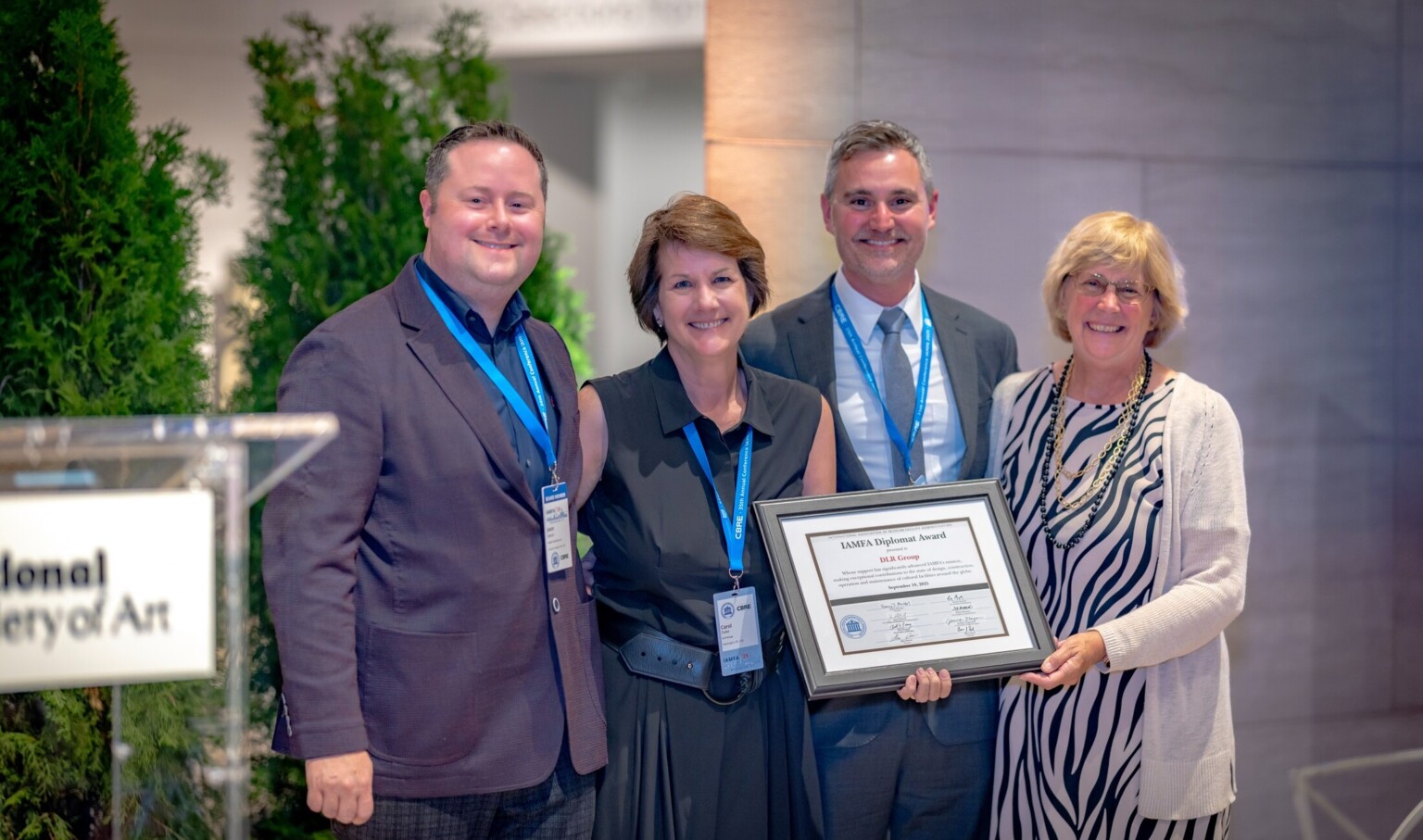 four people standing on a stage to the left of a podium receiving an award. From left: Jason Dittrich, vice president of marketing and communications, IAMFA Board of Directors; Carol Duke, principal and business development leader, Assoc. AIA; Jason Majerus, senior principal and senior engineering leader, PE, CEM, LEED AP; Nancy Bechtol, president, IAMFA Board of Directors