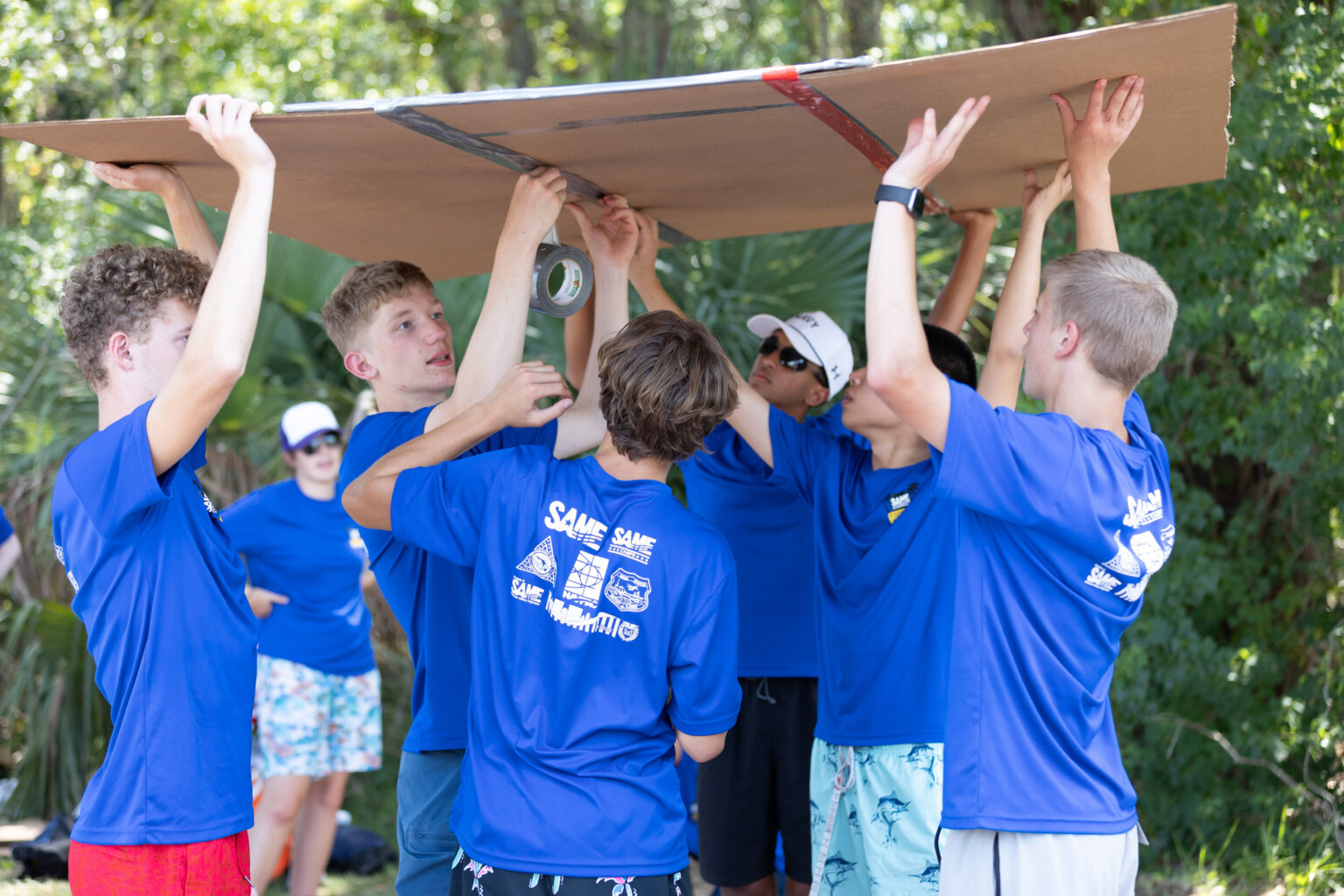 Six STEM campers at Naval Air Station Jacksonville hold a large piece of cardboard over their heads as they apply tape to its underside