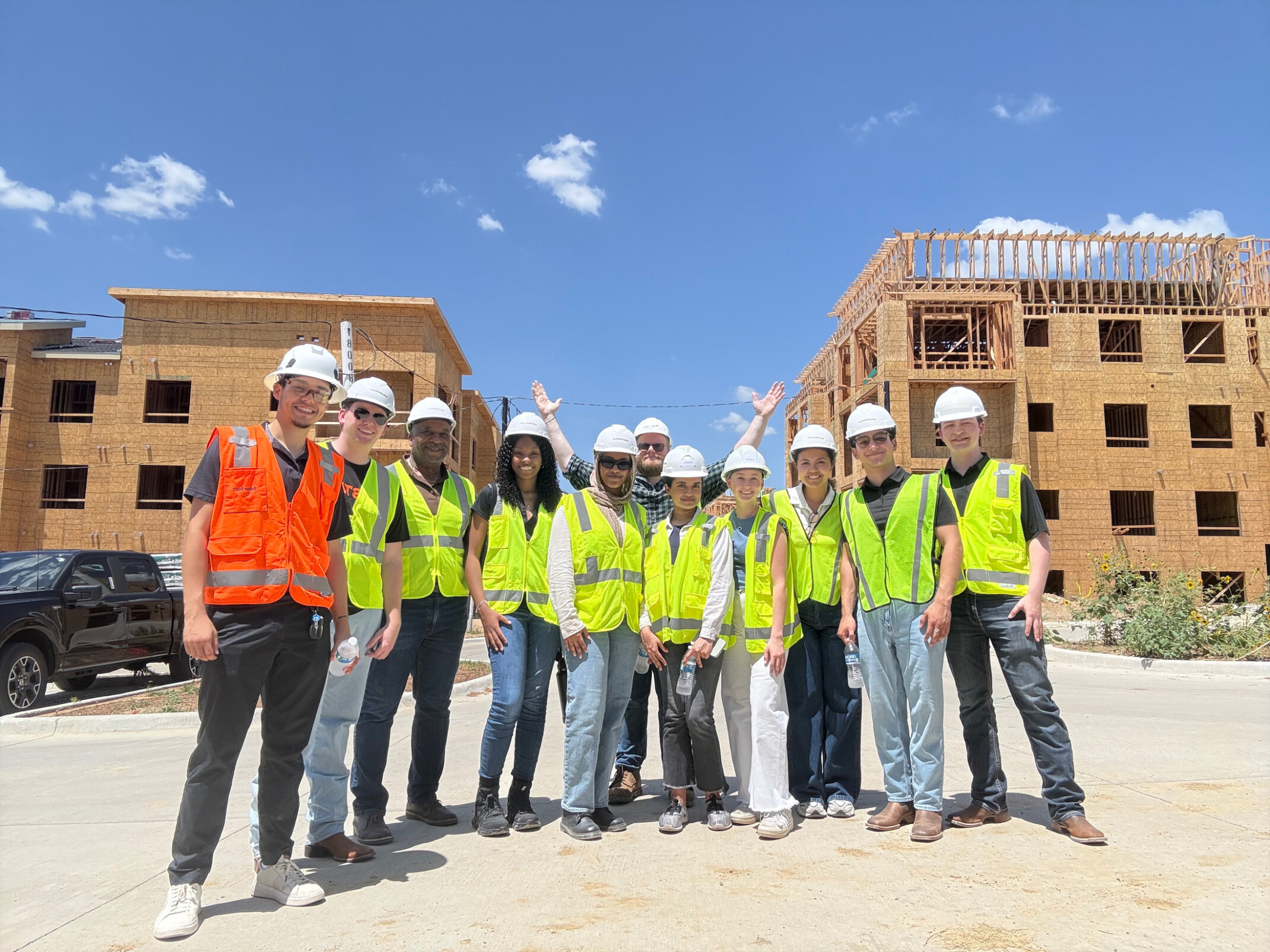 A group of nine college-age people in Hi-Viz vests and hard hats poses in front of a sunny construction site with two partially built multistory structures behind them.