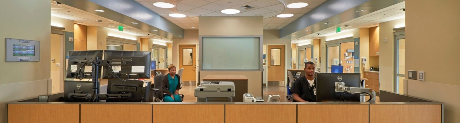 interior front desk of a hospital; facing a warm wood half wall with work stations behind