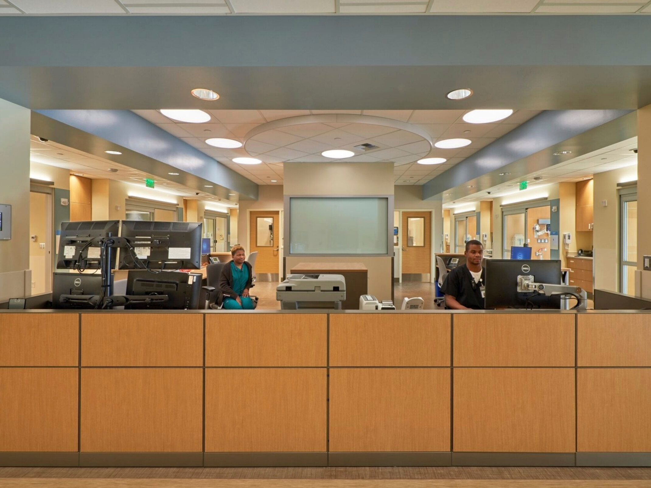 interior front desk of a hospital; facing a warm wood half wall with work stations behind