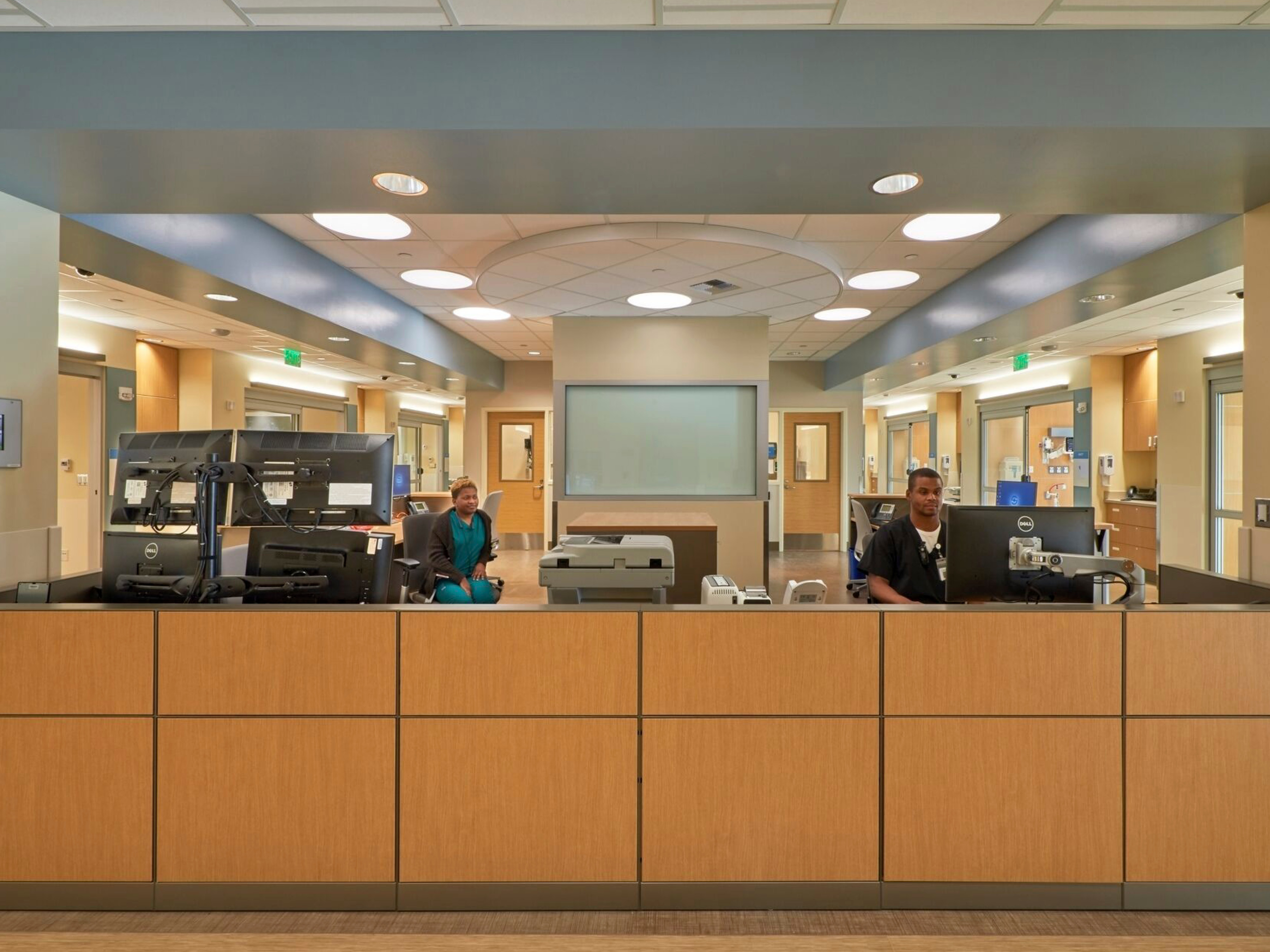 interior front desk of a hospital; facing a warm wood half wall with work stations behind