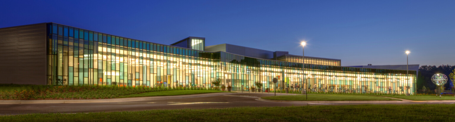nighttime image of a building with colorful floor to ceiling windows with grass and walkways in the foreground