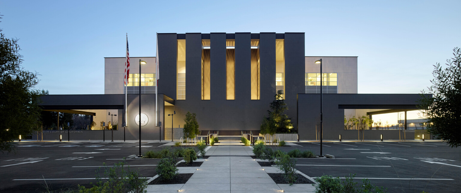 evening view of three story courthouse building designed with dark brick and multistory vertical windows, illuminating the building at night, sidewalk and greenery lead to front door