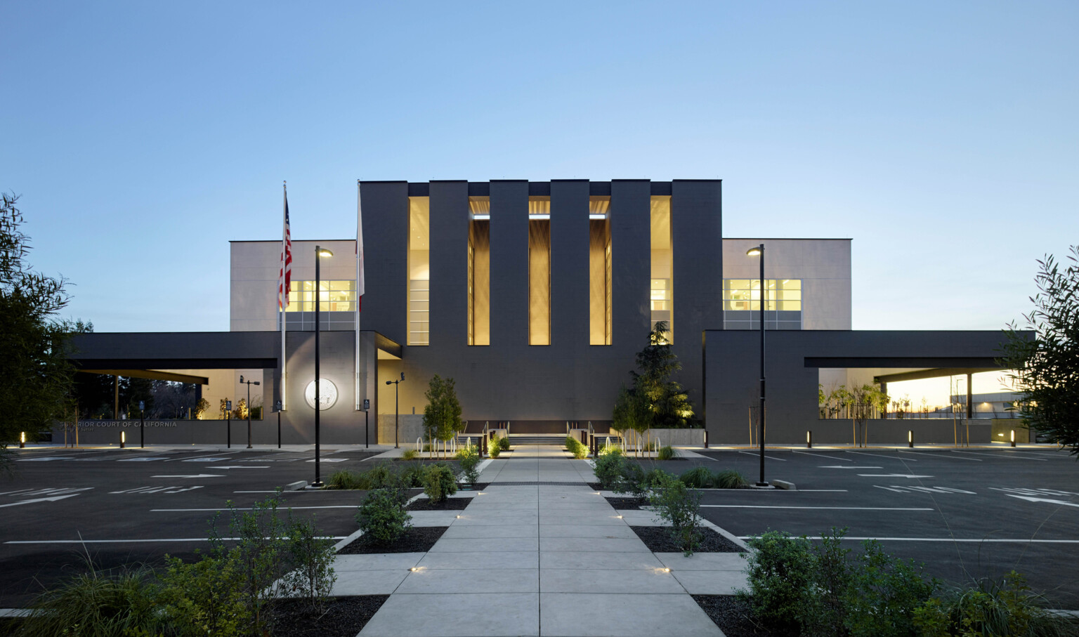 evening view of three story courthouse building designed with dark brick and multistory vertical windows, illuminating the building at night, sidewalk and greenery lead to front door