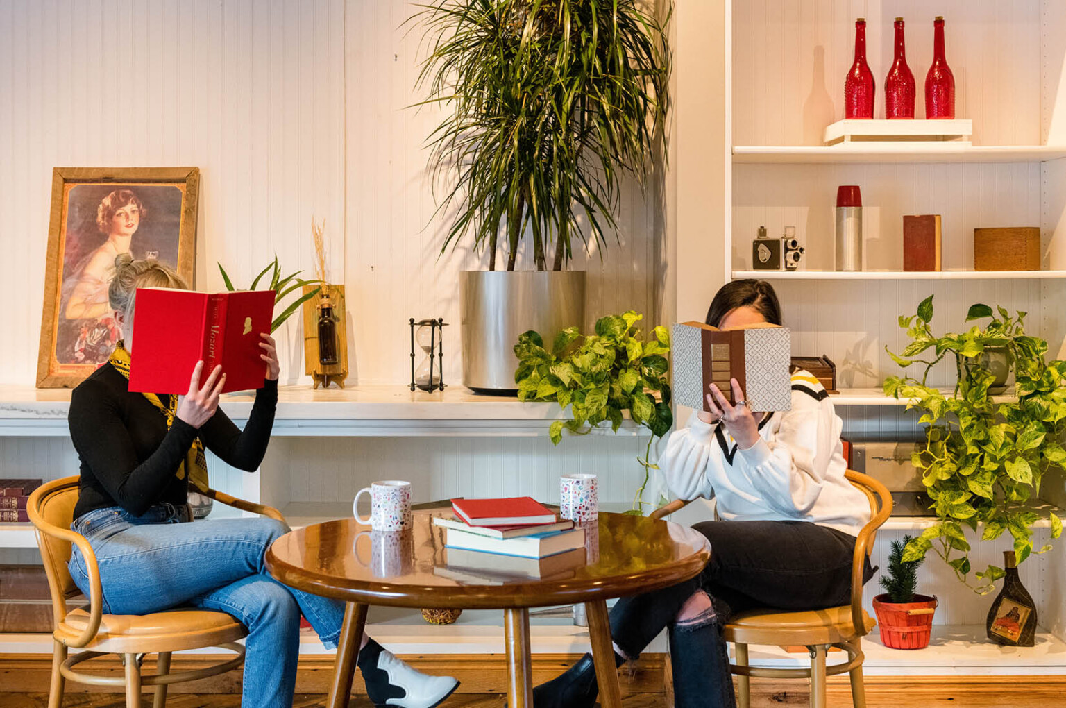 two women sitting at a table reading books and enjoying coffee at a hotel in Colorado. Midcentury modern furniture, plants, painting, bistro table.