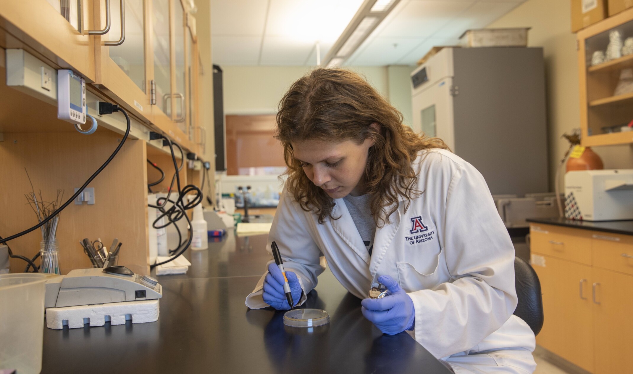 Woman works with specimen and stopwatch at lab table with equipment on black counter with wood cabinetry above and below