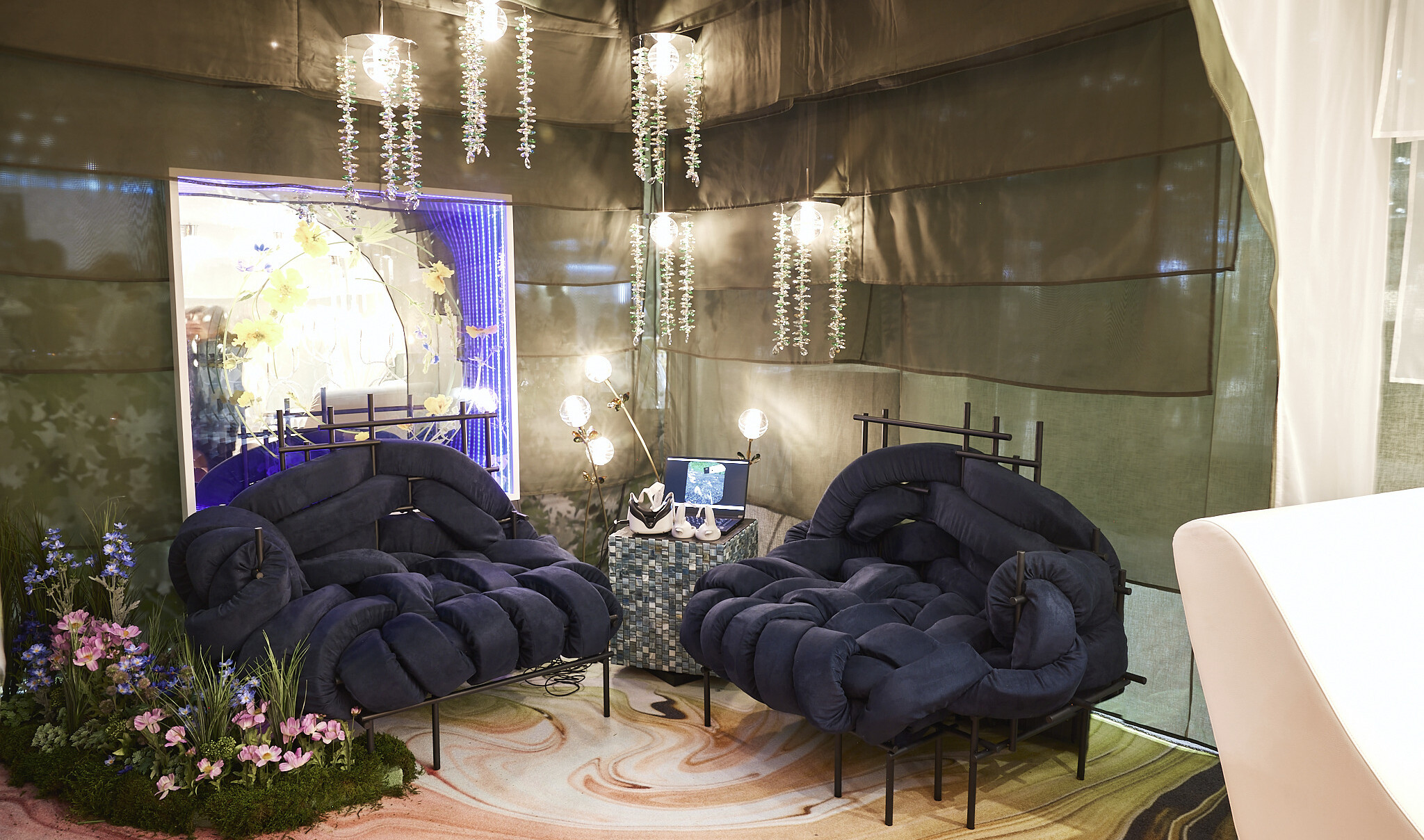 Navy woven cushioned chairs in corner of room with layered fabric partition walls. Greenery on floor, pocket wall accent, and hanging light