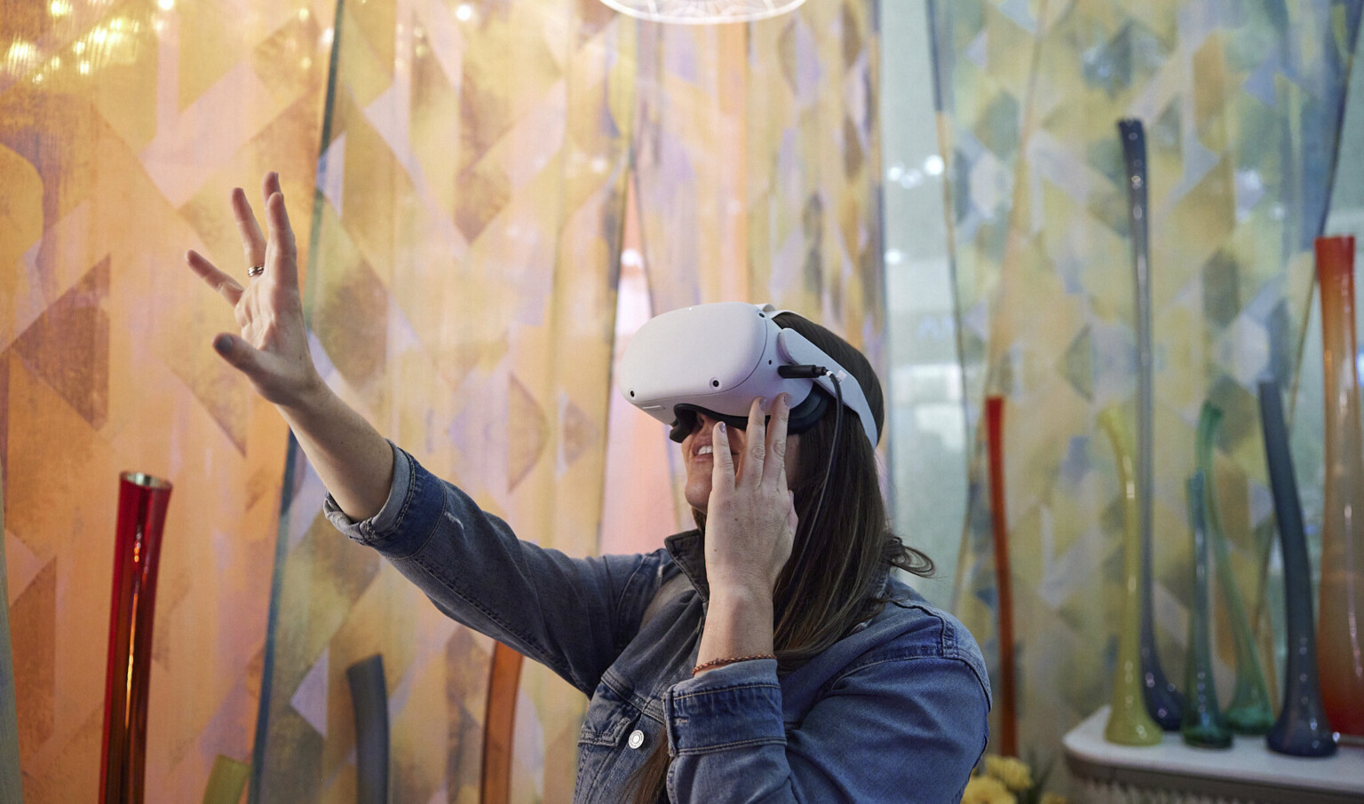 Woman using VR headset reaches out in room with geometric triangular patterned curtains in warm cool tone gradient. Organically shaped vases