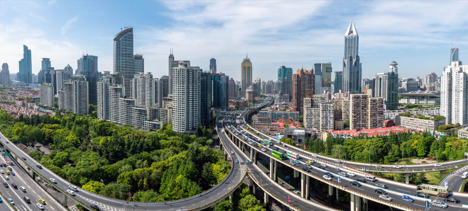 traffic on elevated roads surrounded by trees lead into Shanghai, the city skyline made up of angular high rise buildings