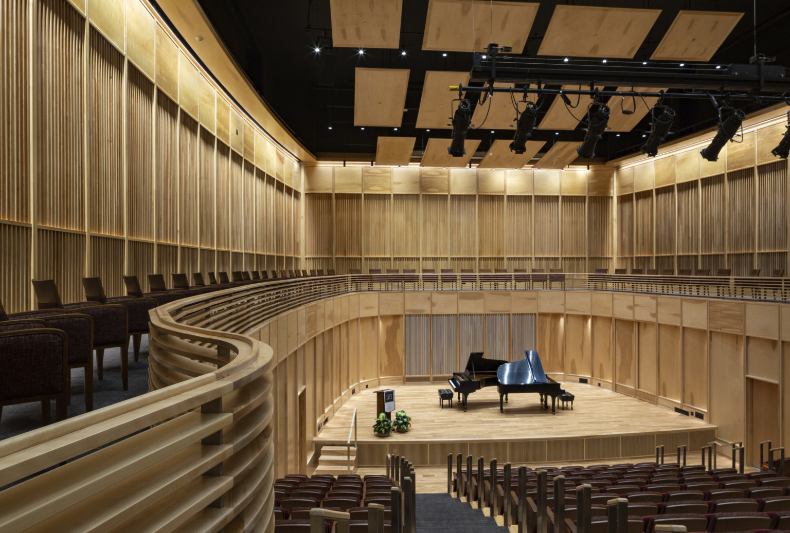 View from the back of wooden wall oval auditorium with piano on stage below wood acoustic panels and balcony behind stage