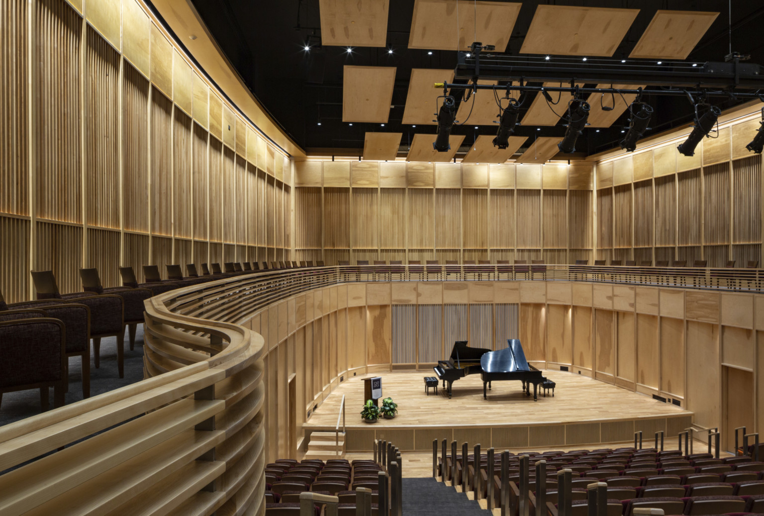 View from the back of wooden wall oval auditorium with piano on stage below wood acoustic panels and balcony behind stage View from the back of wooden wall oval auditorium with piano on stage below wood acoustic panels and balcony behind stage