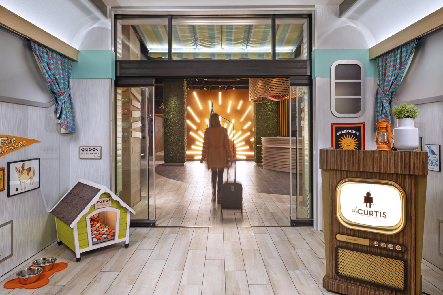 Guest entering through glass double doors towards lit mural on orange wall. Wood reception podium with the Curtis hotel logo Guest entering through glass double doors towards lit mural on orange wall. Wood reception podium with the Curtis hotel logo