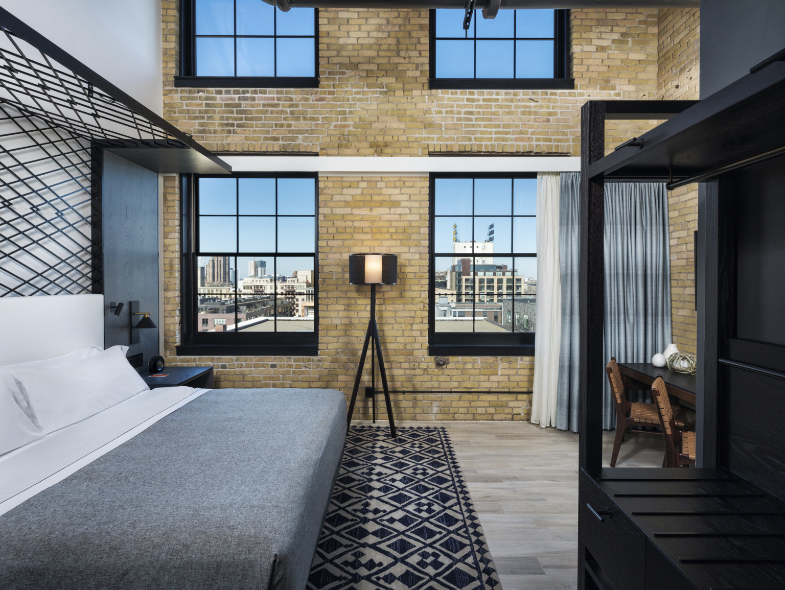 Double height guest room with curtains from lower windows. Dark metallic canopy over bed, left, wood dresser and table, right