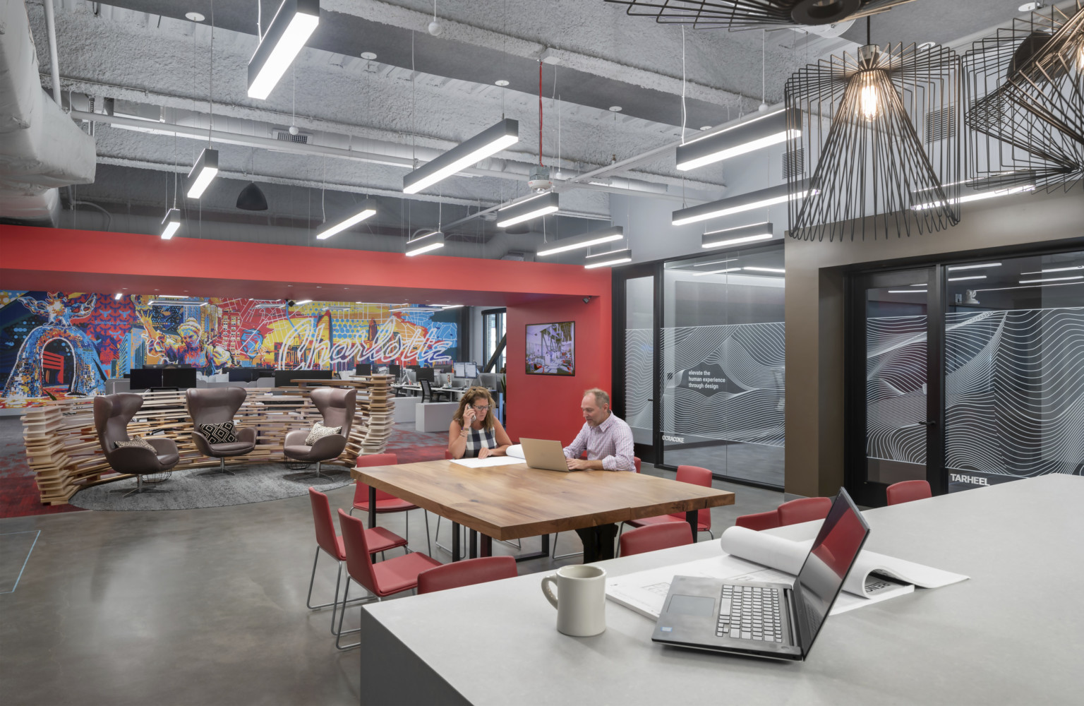 two people sit at a commons table with red chairs in the charlotte office with large mural on red wall behind them