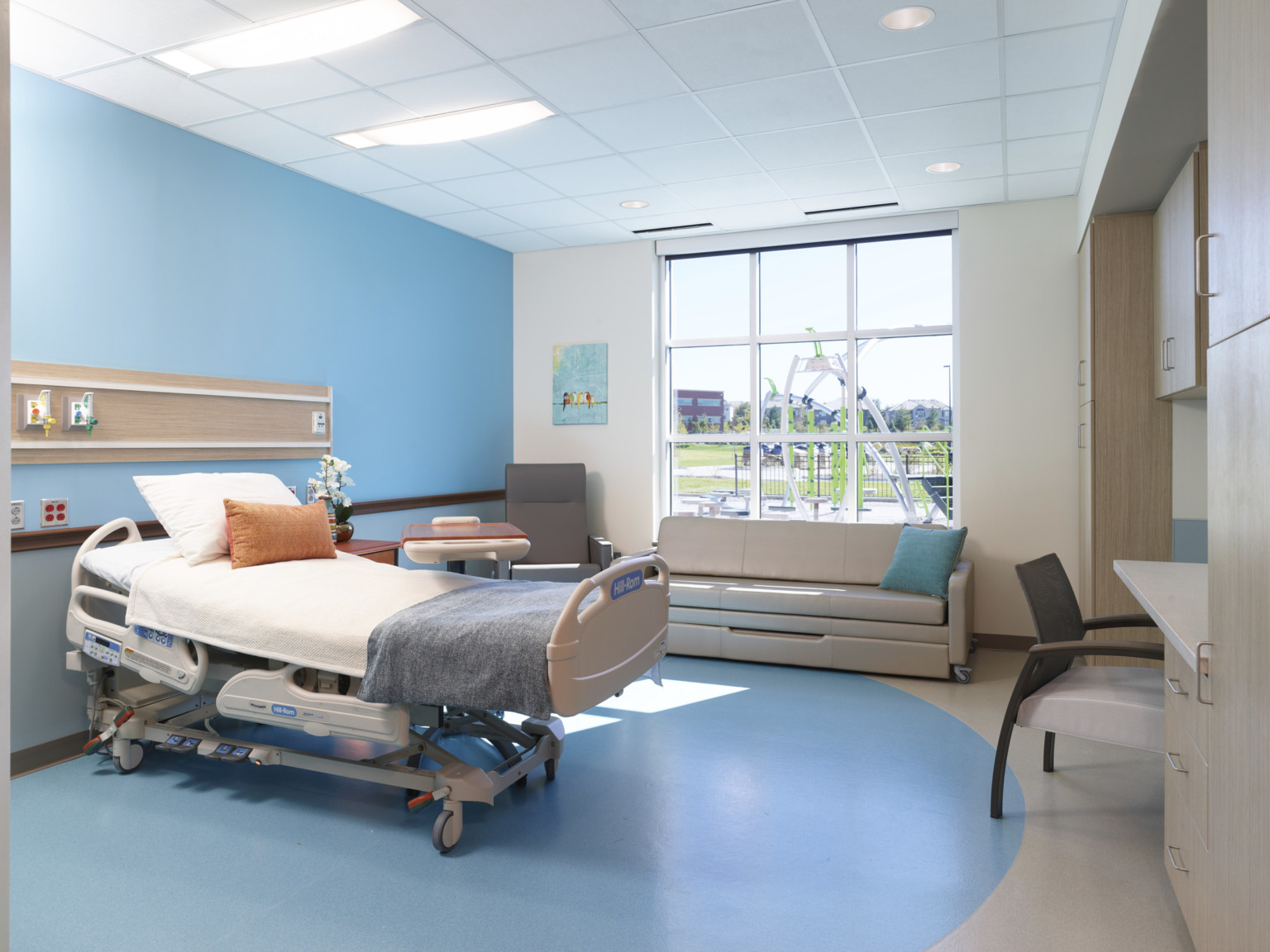 Hospital bed faces desk against blue wall connected to blue semi circle on grey floor. Couch faces bed in front of window