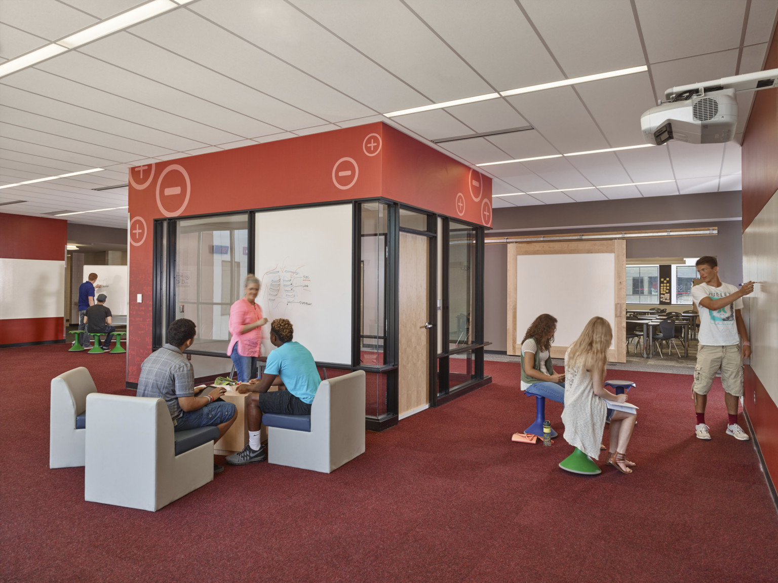 Nontraditional seating areas in front of white boards in room with red walls and carpets. Glass room with wood door, center
