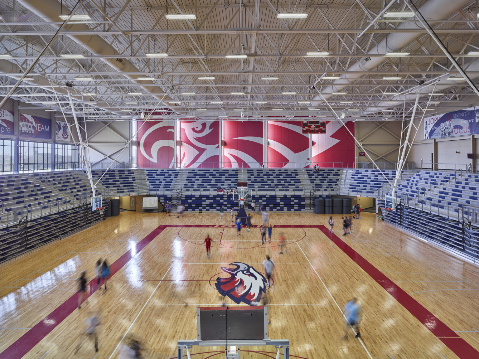 Double height wood floor gymnasium. Blue and white bleachers at sides with retracted lower deck. Eagle mural on back wall