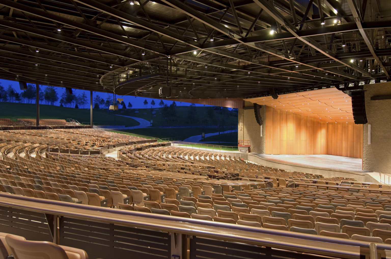 Light brown seats under black amphitheater roof facing wood paneled stage with rounded panel ceiling and recessed lights