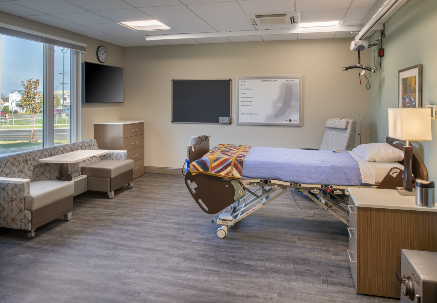 hospital room with patient bed on wheels integrated track ceiling wood floor and neutral walls. exterior wall has tall window