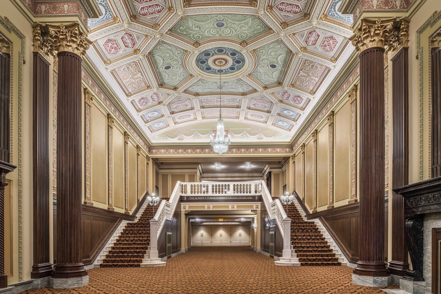 Grand staircase carpeted beneath ornate multicolored coffered ceiling with chandelier framed by ribbed pillars