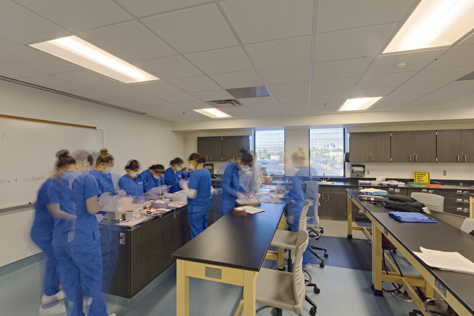 students in blue scrubs working at a lab station with a whiteboard behind them and daylight shining in from the far exterior windows
