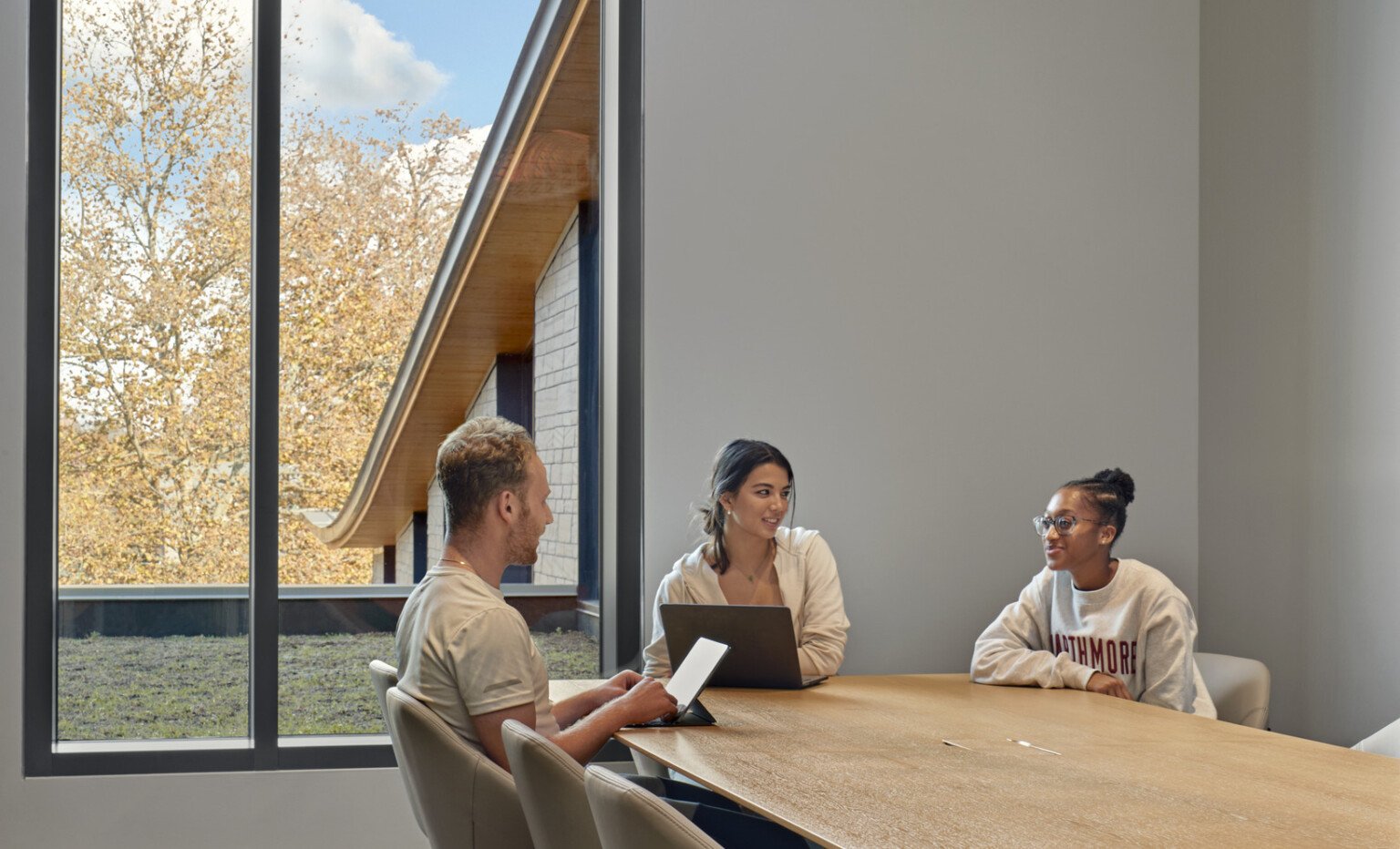 Long wood table with chairs with tan upholstery in tall ceiling room with large windows. View to sloping roof and overhang