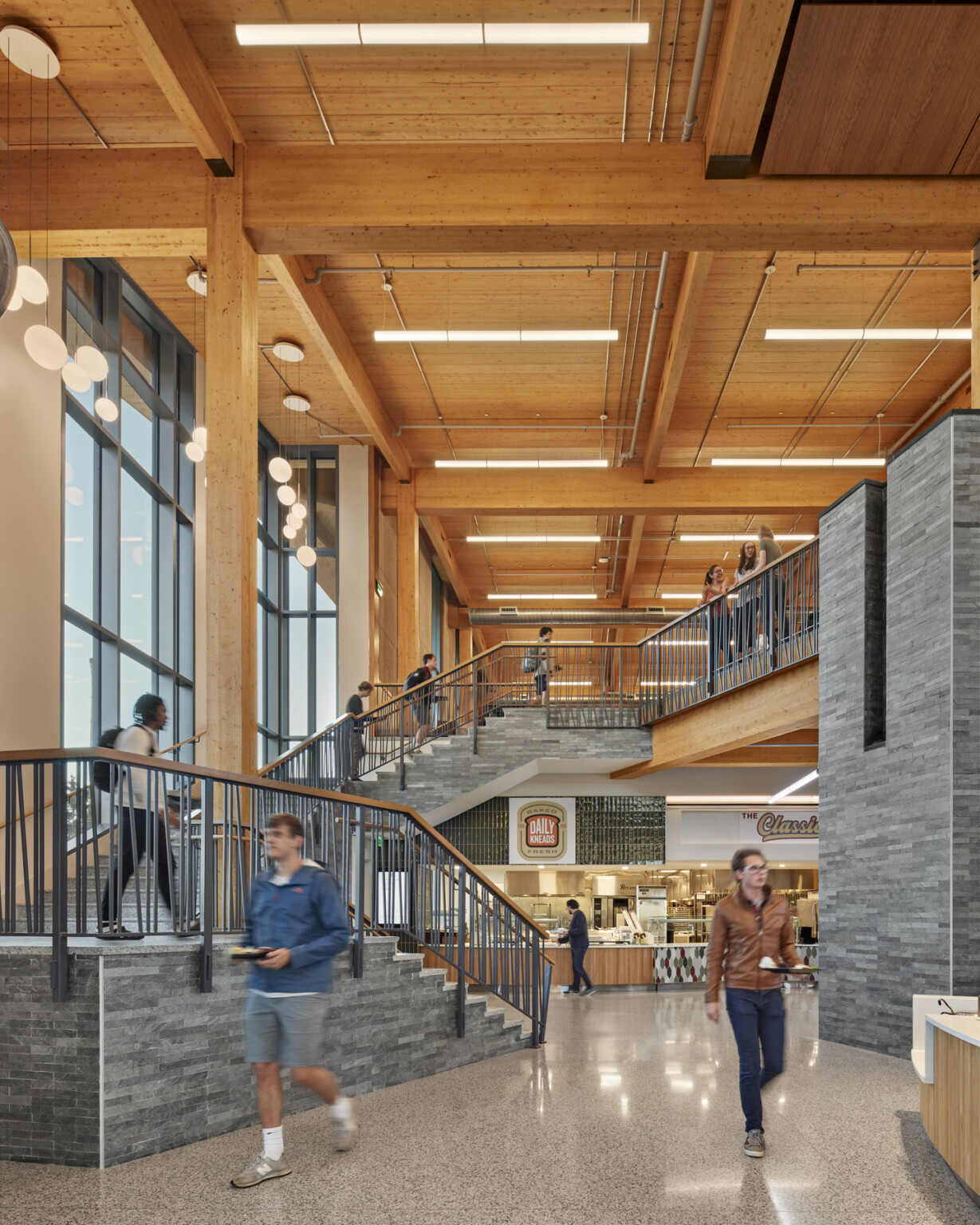 Double height atrium with exposed wood mass timber beams, columns, and ceiling. Grey accents. Dining options along walls, balcony above