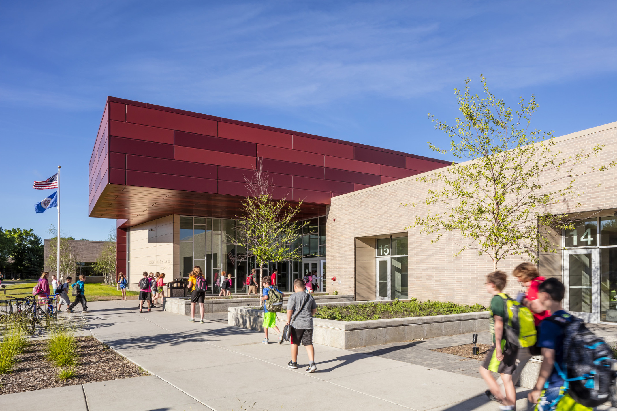 Jordan Middle School exterior entrance with floor to ceiling windows around doors, beige brick walls, and a dark red overhang Jordan Middle School exterior entrance with floor to ceiling windows around doors, beige brick walls, and a dark red overhang