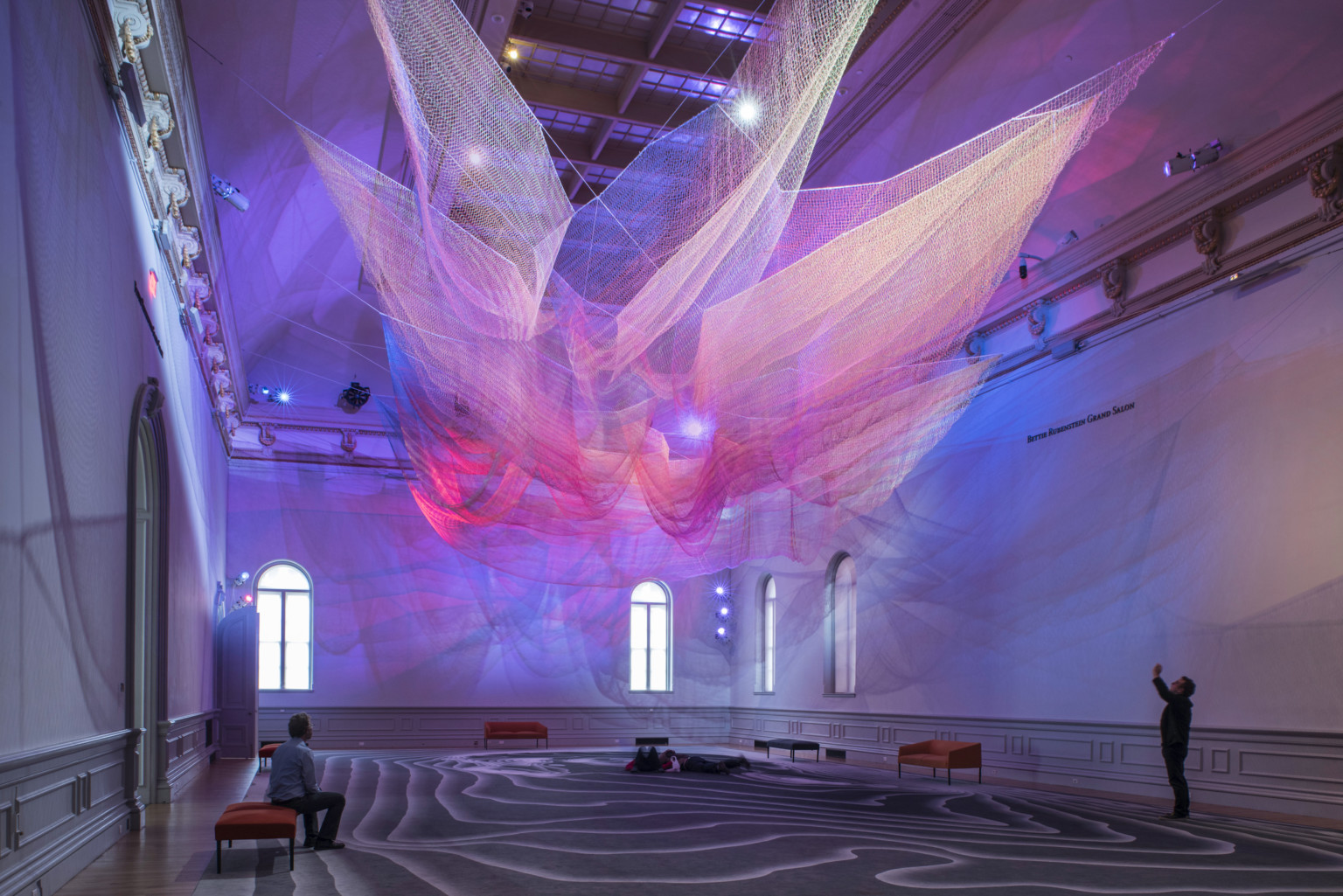 Colorful abstract art installation with pink and purple light on layers of sheer netting with shadows and light on white wall